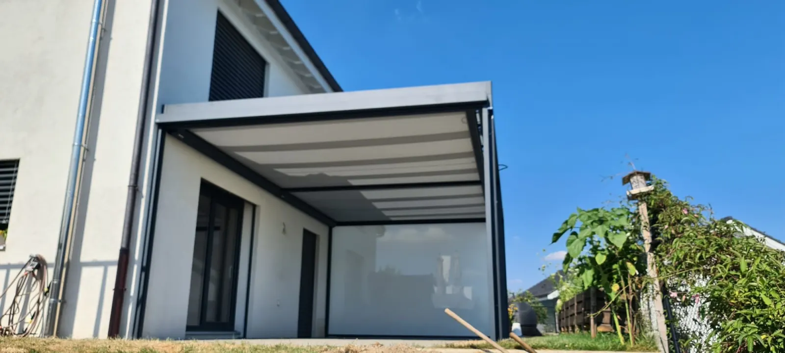 Terrasse protégée par une structure aluminium et un store vertical clair, posé devant une maison contemporaine.